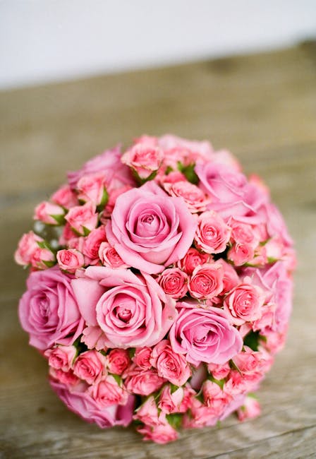 A bouquet arrangement of fresh pink roses and smaller pink spray roses, closely grouped into a dense, rounded cluster. The roses have fully opened, displaying delicate, velvety petals with subtle vari